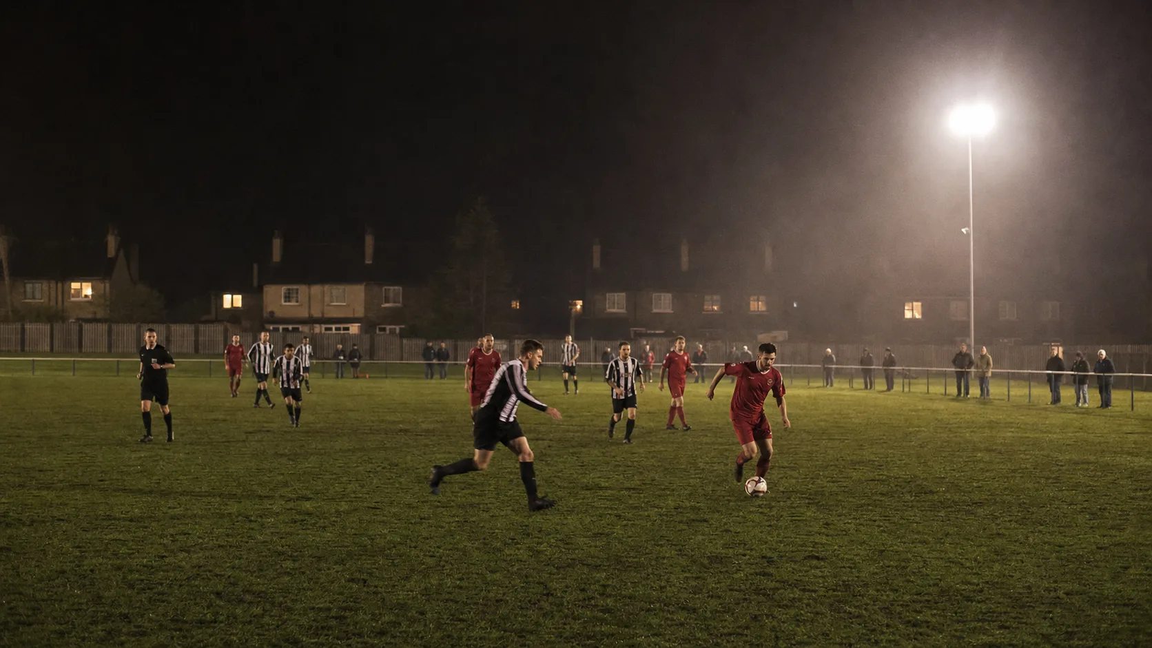 Community football players cross a damp neighborhood pitch under modest floodlights.
