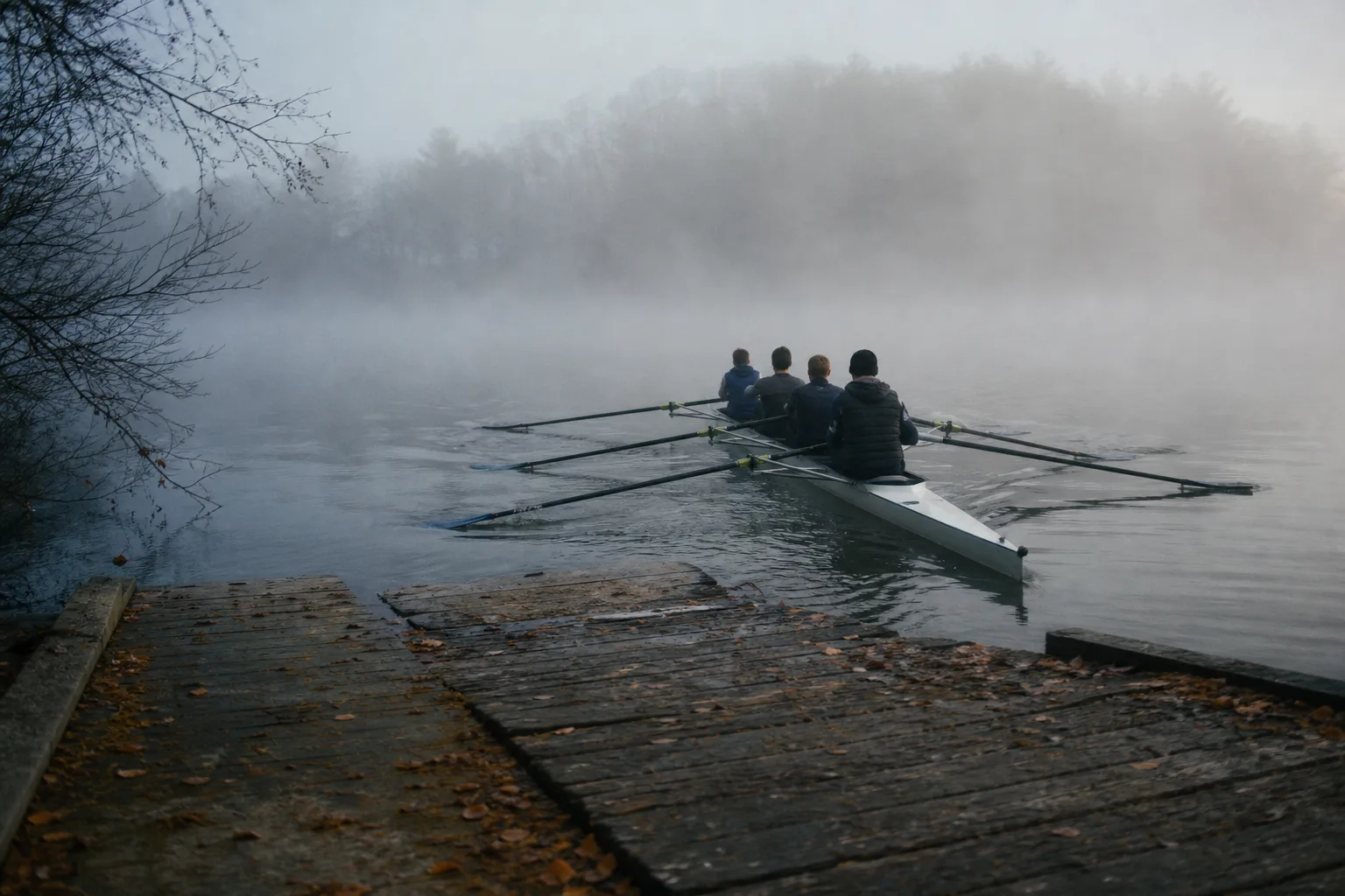 Four community rowers push a narrow shell away from a timber landing into morning fog.
