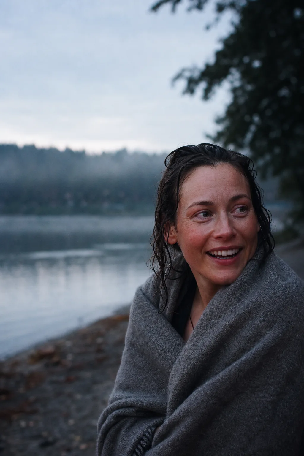 An open-water swimmer wrapped in a grey blanket smiles toward friends after leaving the lake.